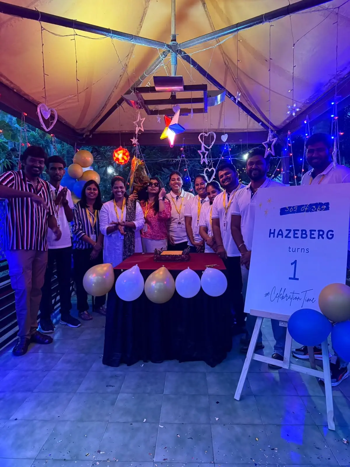 The team around a cake under party lights, beside a sign that reads Hazeberg turns 1